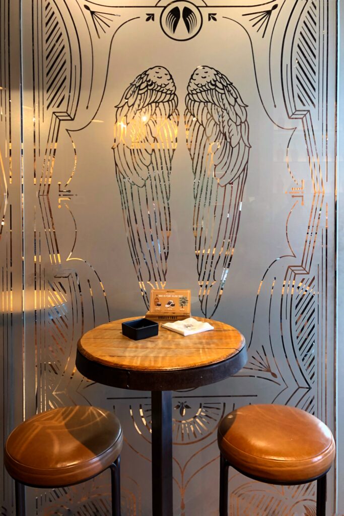 Angel’s Envy Distillery tasting room in Louisville, Kentucky with an Art Deco–style frosted glass panel etched with angel wings behind a small round table and leather bar stools—an aesthetic stop on the Kentucky Bourbon Trail.