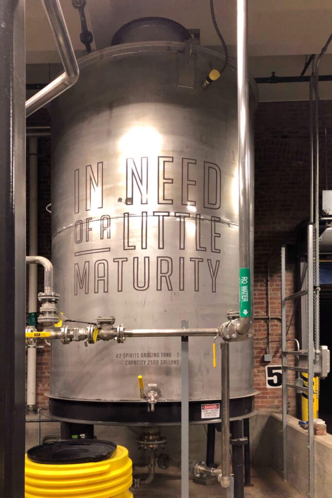 Angel’s Envy Distillery production area in Louisville, Kentucky showing a large stainless maturity tank labeled “In Need of a Little Maturity,” with pipes and valves against exposed brick—Kentucky Bourbon Trail tour stop.