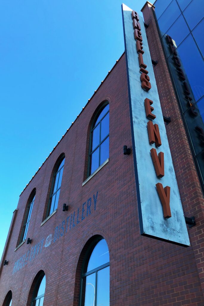 Exterior of Angel’s Envy Distillery in Louisville, Kentucky with a tall vertical “Angel’s Envy” sign on a red brick building under a clear blue sky—an iconic stop on the Kentucky Bourbon Trail.