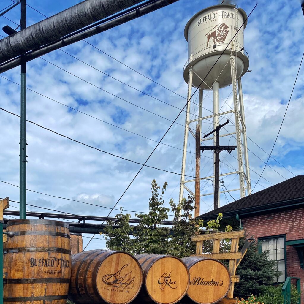Buffalo Trace Distillery in Frankfort, Kentucky with the iconic water tower and bison logo rising above stacked bourbon barrels and brick warehouses under a blue sky—classic stop on the Kentucky Bourbon Trail.