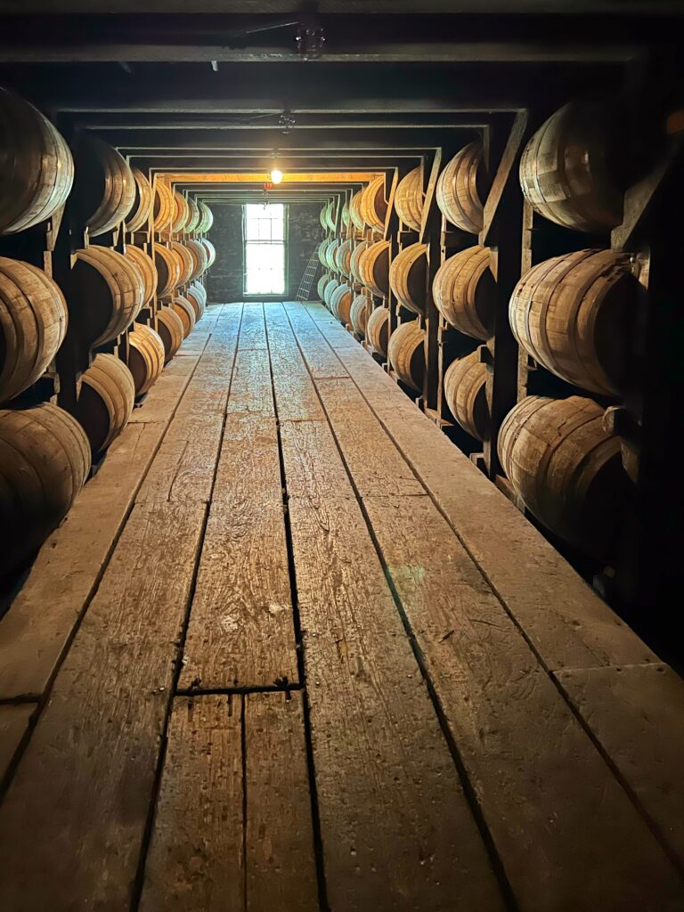 Dimly lit bourbon rickhouse on the Kentucky Bourbon Trail with rows of aging oak barrels stacked on wooden racks along a narrow plank walkway leading to a bright window at the end of the warehouse.