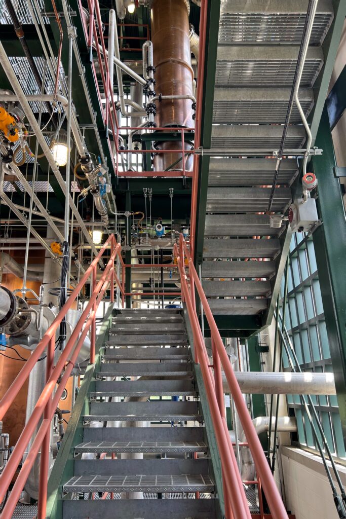 Buffalo Trace Distillery Tour - Metal staircase inside a Kentucky Bourbon Trail distillery with red railings, green steel beams, exposed pipes, and a tall copper still column rising beside factory windows—industrial-chic bourbon production tour.