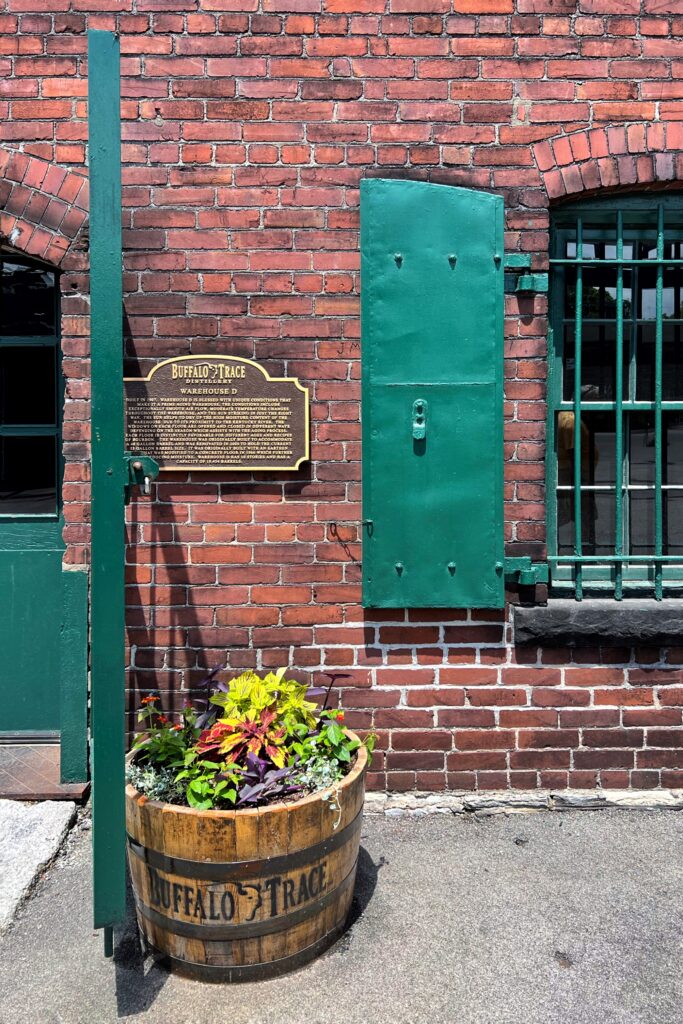 Buffalo Trace Distillery Warehouse D sign on a red brick building in Frankfort, Kentucky, with green shutters and a bourbon barrel planter—an aesthetic stop on the Kentucky Bourbon Trail.