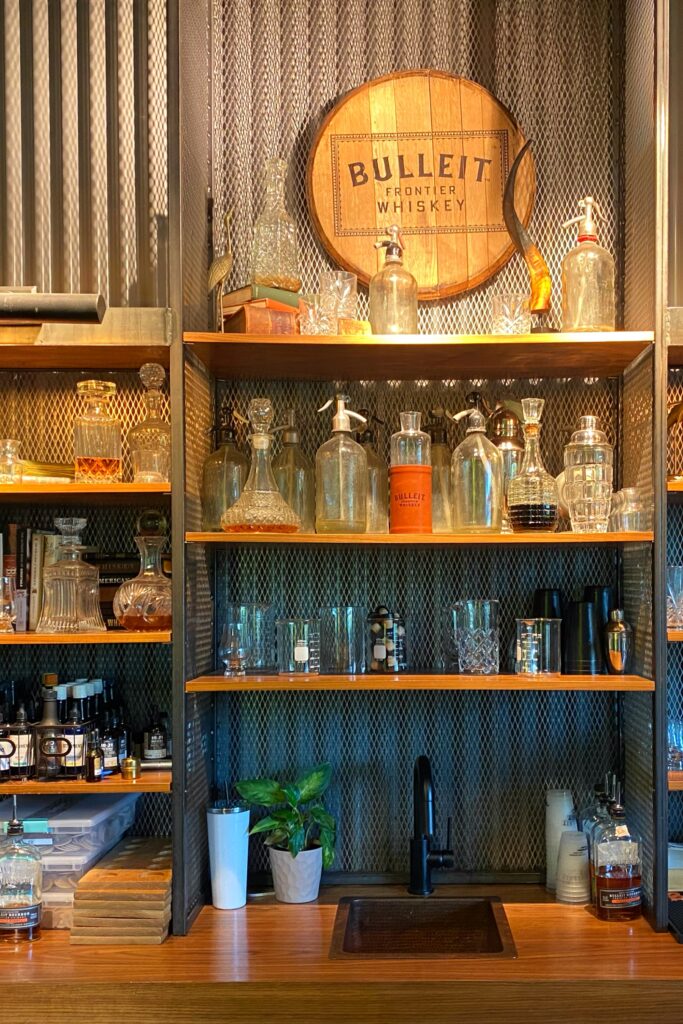 Back bar display at Bulleit Frontier Whiskey with vintage glassware, siphon bottles, etched barware, warm wood shelving, black mesh paneling, and a branded Bulleit barrel plaque.