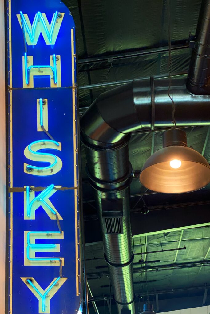 Blue neon whiskey sign at Bulleit Frontier Whiskey with exposed black ductwork, a metal pendant light, and an industrial-style ceiling inside the visitor center.