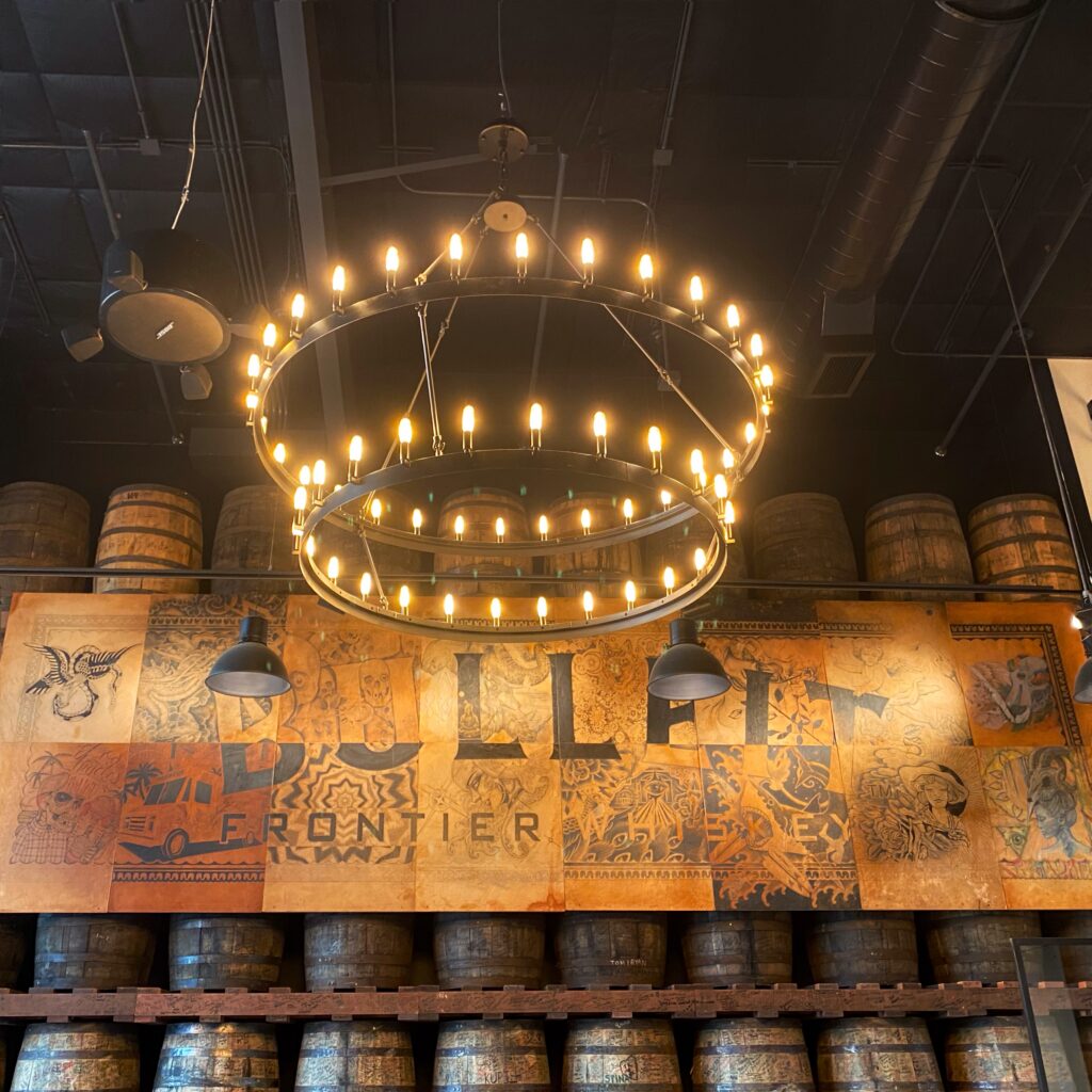 Bulleit Frontier Whiskey interior with stacked bourbon barrels, a large illustrated Bulleit mural, industrial pendant lights, and a dramatic double-ring chandelier. Part of the Aesthetic Bourbon Trail Travel Guide