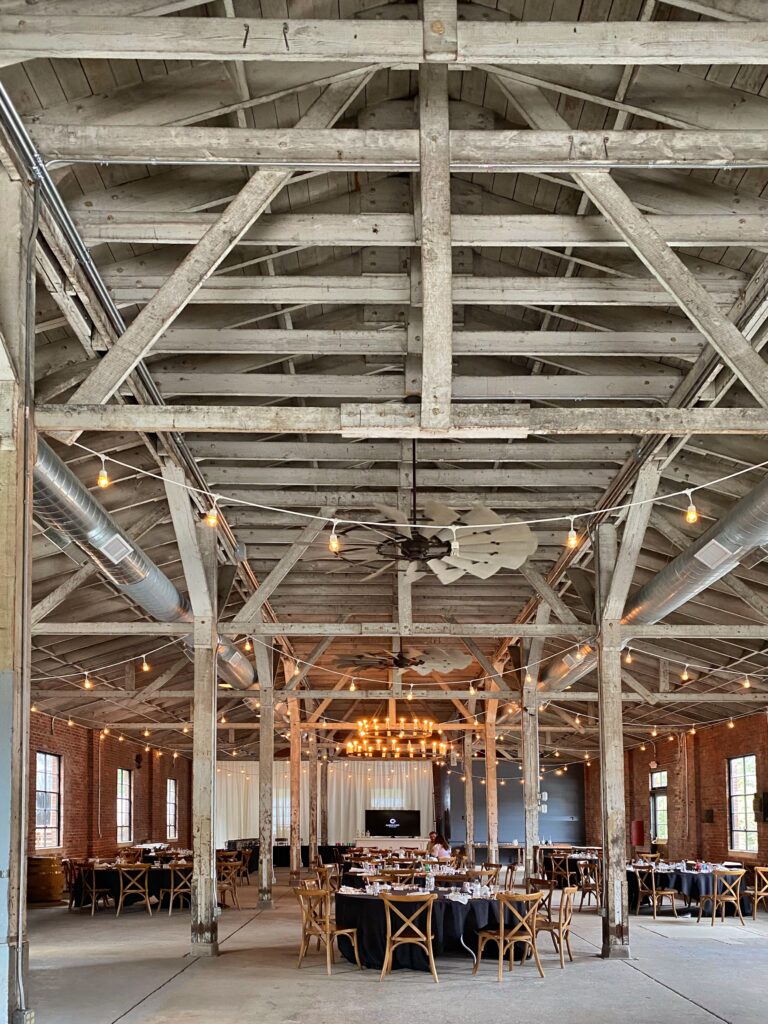 Rustic event barn at Castle & Key Distillery in Frankfort, Kentucky with weathered white timber trusses, exposed brick walls, string lights, and banquet tables set for a private dinner—an aesthetic stop on the Kentucky Bourbon Trail.