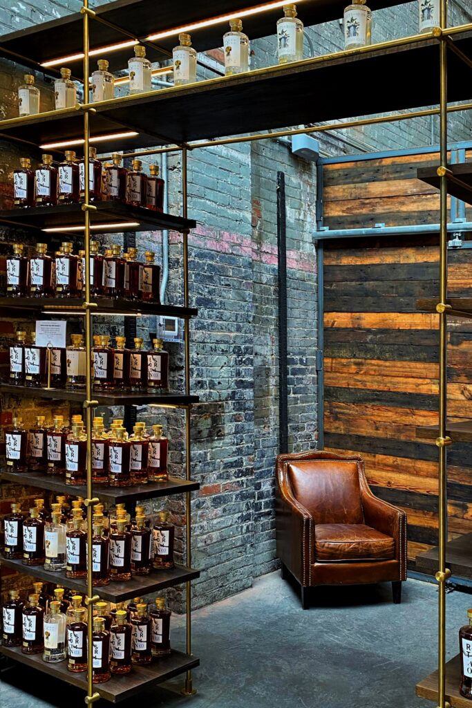 Castle and Key - aesthetic bourbon distillery gift shop on the Kentucky Bourbon Trail with brass shelving lined with whiskey bottles, exposed brick walls, a rustic wood door, and a cognac leather armchair—industrial-chic tasting room vibe in Kentucky.