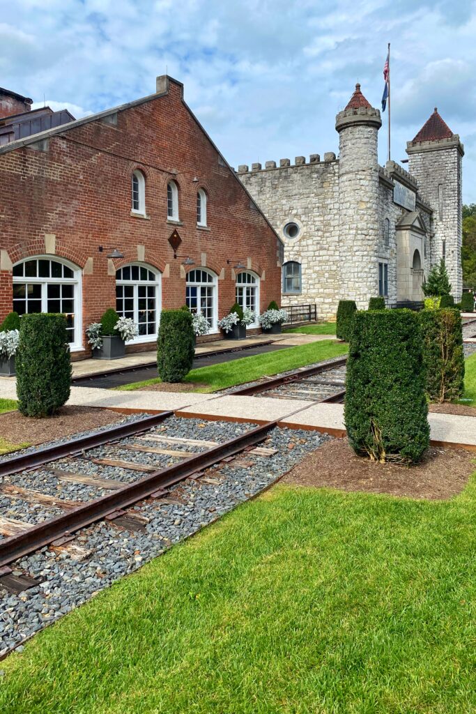 Castle & Key Distillery grounds at the historic Old Taylor Distillery in Frankfort, Kentucky, with a red brick building, castle-like stone towers, manicured hedges, and rail tracks crossing the lawn—an iconic Kentucky Bourbon Trail stop.