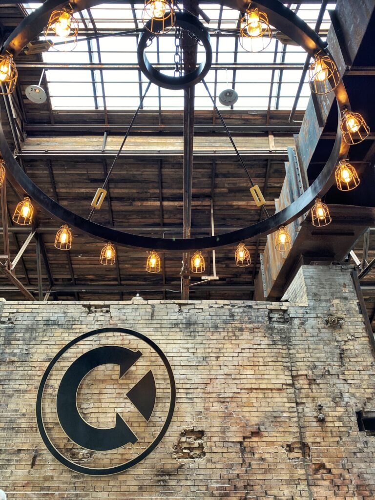 Castle & Key Distillery in Frankfort, Kentucky showing the CK logo on exposed brick beneath a huge circular ring chandelier with caged Edison bulbs and skylights in a restored warehouse—an aesthetic Kentucky Bourbon Trail stop.
