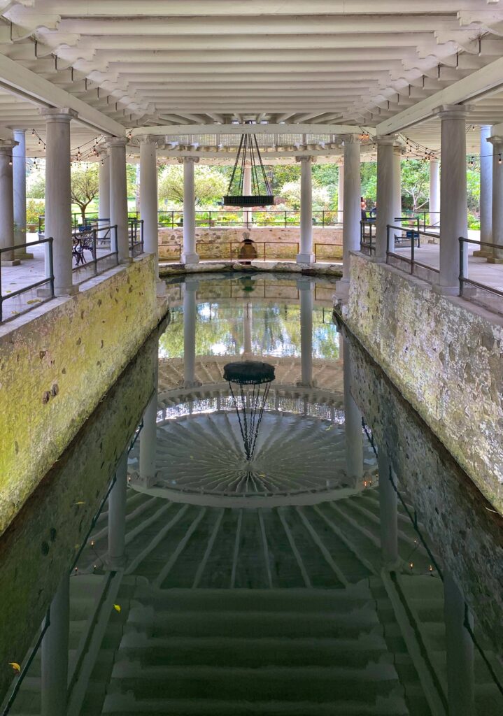 Covered springhouse at the Castle and Key Distillery on the Kentucky Bourbon Trail with a white columned pavilion and calm reflection pool mirroring the circular roof structure, string lights, and stone-lined water channel—an aesthetic distillery stop in Kentucky.