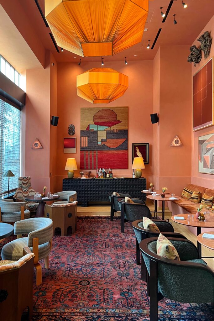 Dahlia Lounge, designed by Kelly Wearstler, at the Proper Hotel in Los Angeles with blush-pink walls, oversized orange pendant lanterns, a patterned red rug, and low lounge seating around small cocktail tables.