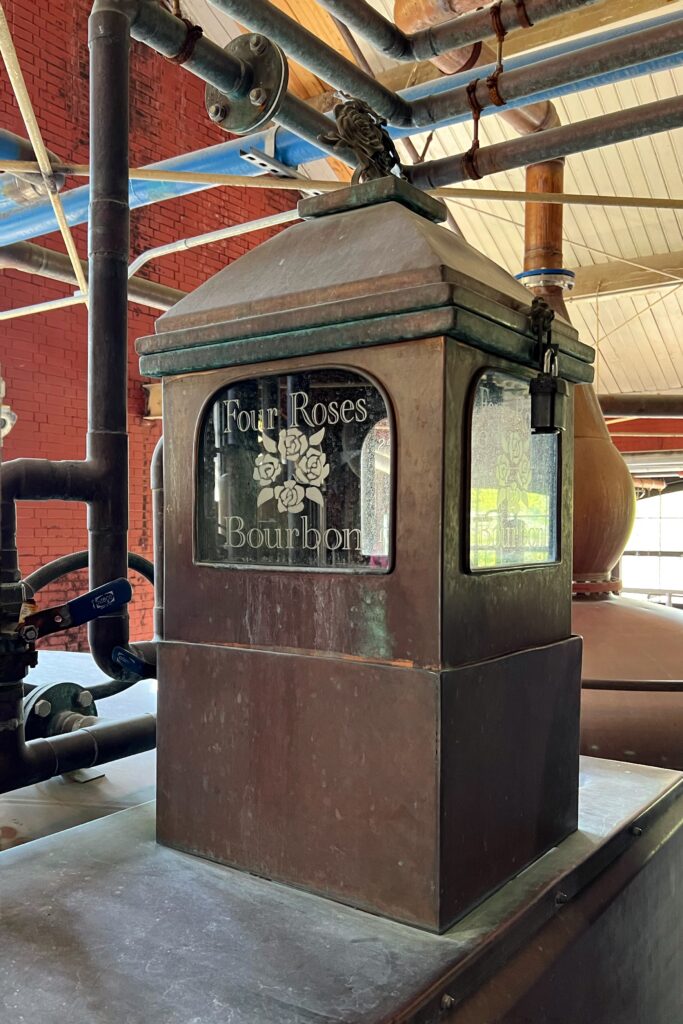 Aged copper equipment detail at Four Roses Distillery in Kentucky, surrounded by industrial piping, red brick walls, and the etched Four Roses Bourbon logo.