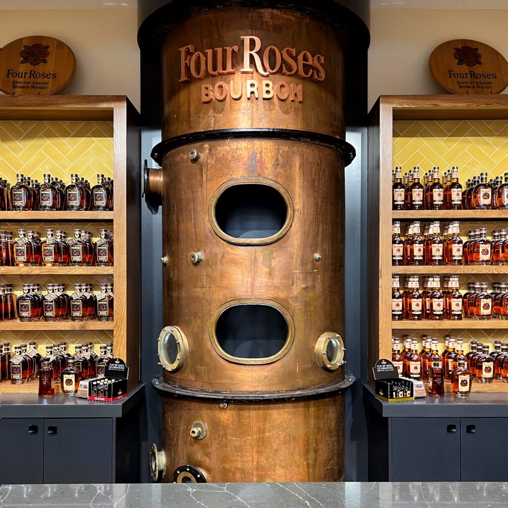 Copper still display at Four Roses Distillery in Kentucky, framed by shelves of Four Roses Bourbon bottles and warm herringbone tile in the tasting room.