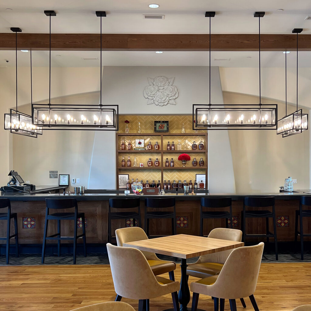 Tasting room bar at Four Roses Distillery on the Kentucky Bourbon Trail, featuring a long wood bar, black barstools, geometric pendant lights, cream stucco walls, and warm wood flooring.