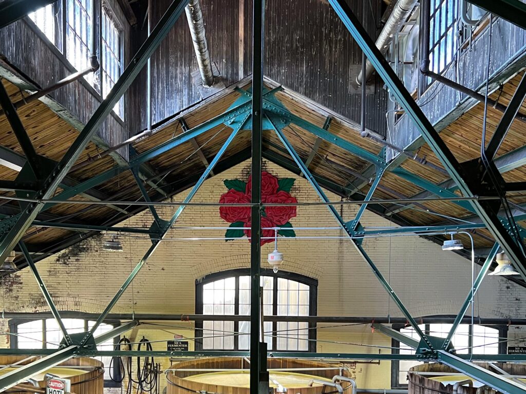 Interior of Four Roses Distillery in Lawrenceburg, Kentucky featuring teal steel trusses, weathered wood rafters, tall factory windows, and a painted red rose mural above brick arches—an aesthetic stop on the Kentucky Bourbon Trail.