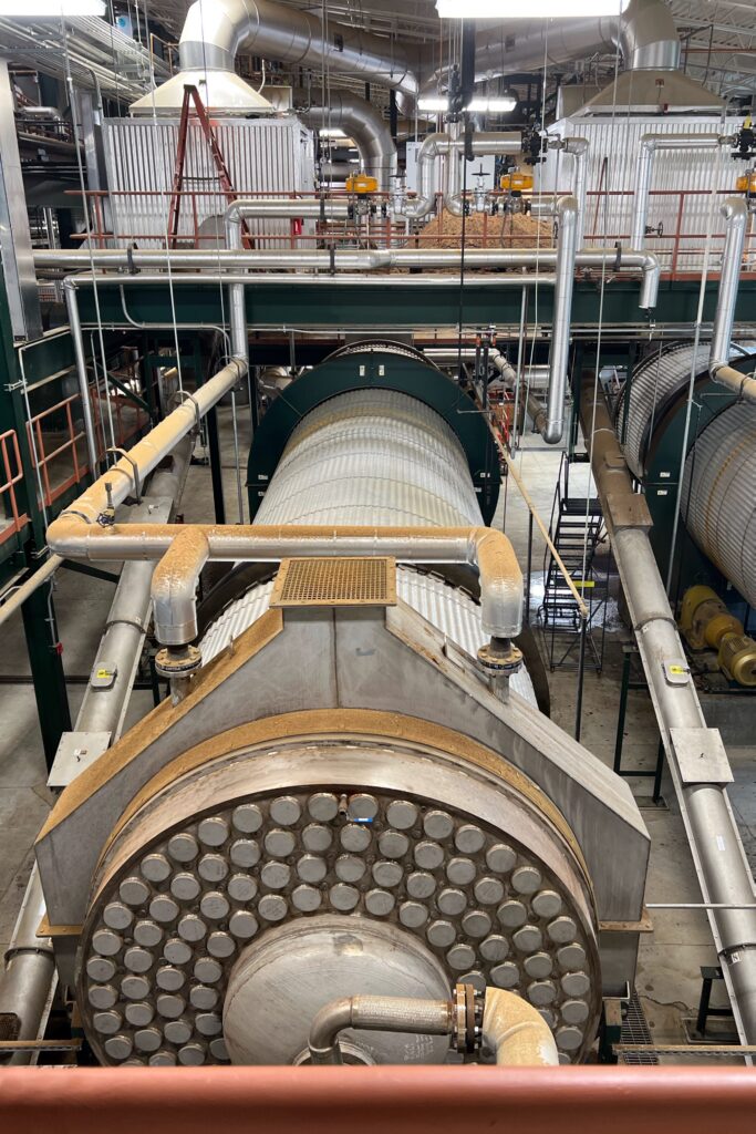 Buffalo Trace industrial bourbon distillery production room on the Kentucky Bourbon Trail with a large stainless-steel still column, silver ductwork and pipes, and grain handling equipment viewed from an overhead catwalk.