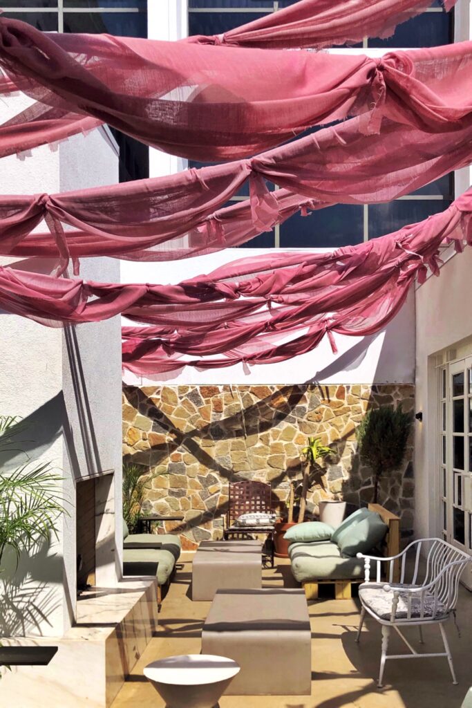 Courtyard patio at The LINE Hotel Austin with billowy blush-pink fabric canopies draped overhead, casting shadows on lounge seating and a stone accent wall in bright sunlight.