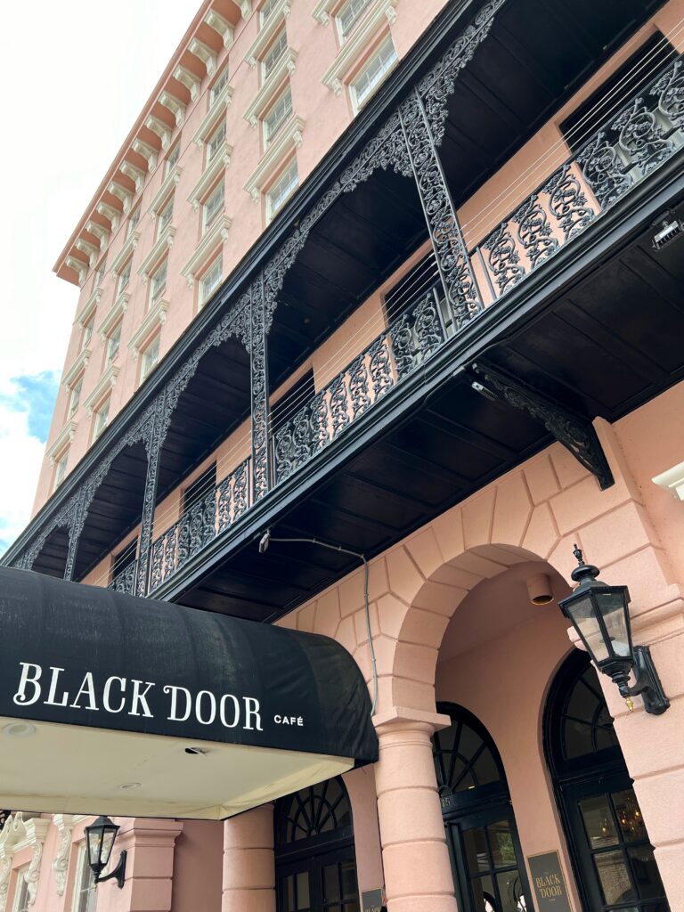Blush-pink stucco façade of the Mills House Hotel in Charleston, South Carolina, with black wrought-iron wraparound balconies above the Black Door Café coffee shop awning and arched entryway.