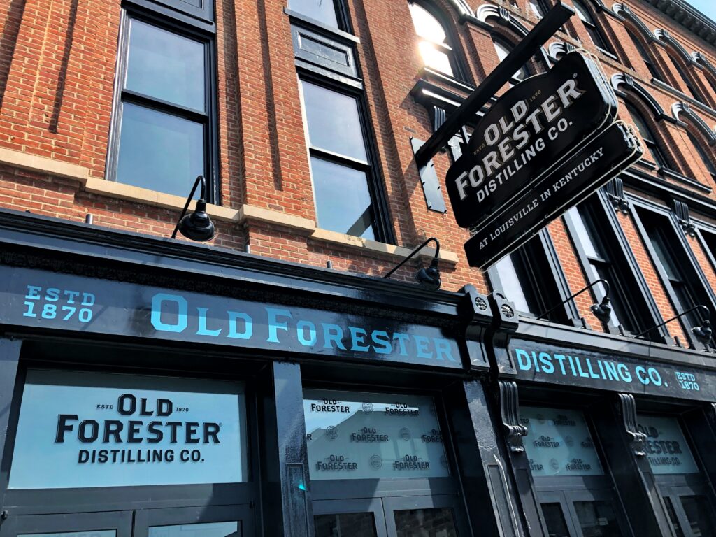 Exterior of Old Forester Distilling Co. in Louisville, Kentucky, with a historic red brick facade, black painted storefront, and hanging distillery sign on Whiskey Row.