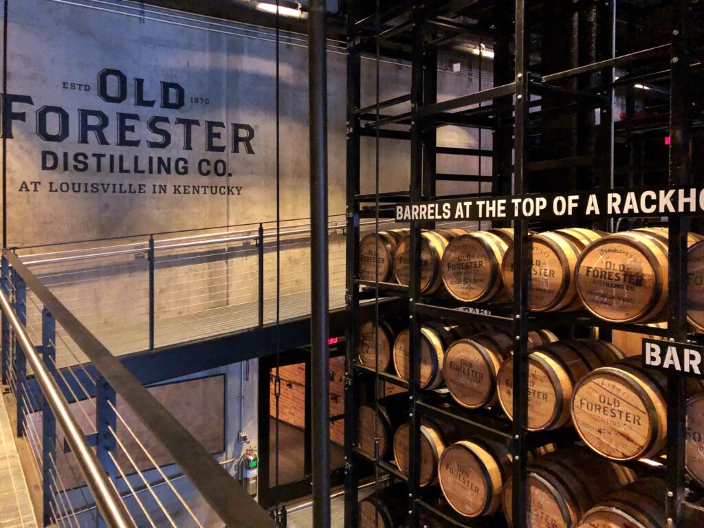 Rackhouse-style barrel display at Old Forester Distilling Co. in Louisville, Kentucky, with stacked bourbon barrels, black steel rack framing, an elevated catwalk, and large industrial wall branding.