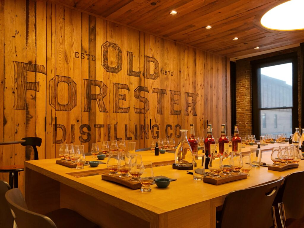 Tasting room at Old Forester Distilling Co. in Louisville, Kentucky, with wood-paneled walls, communal tasting tables, bourbon flight glasses, and a large branded wall graphic.