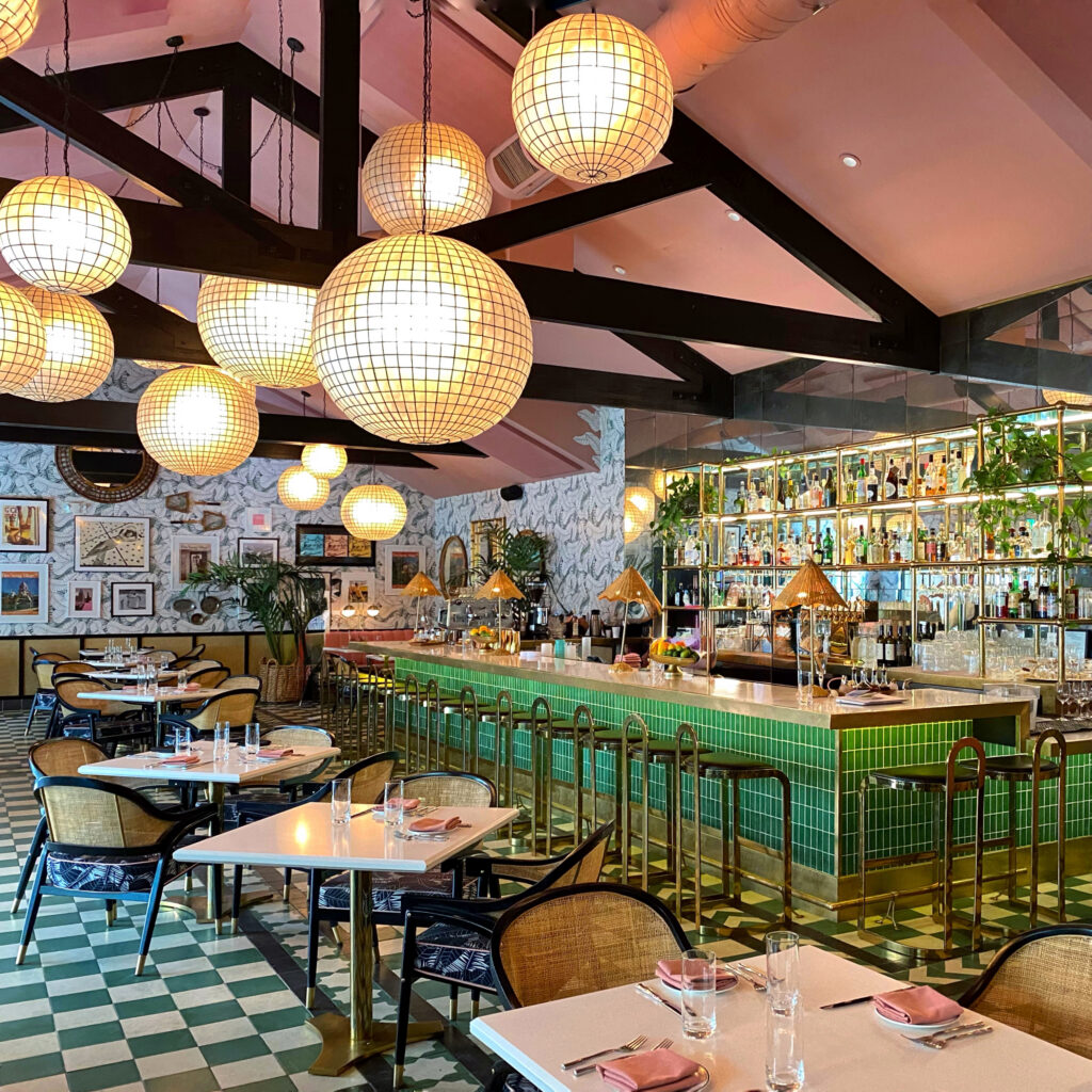 Interior of Pink Cabana at Indian Wells near Palm Springs featuring a blush-pink ceiling with black beams, oversized globe pendant lights, a green-tiled bar, and white tables set with pink napkins.