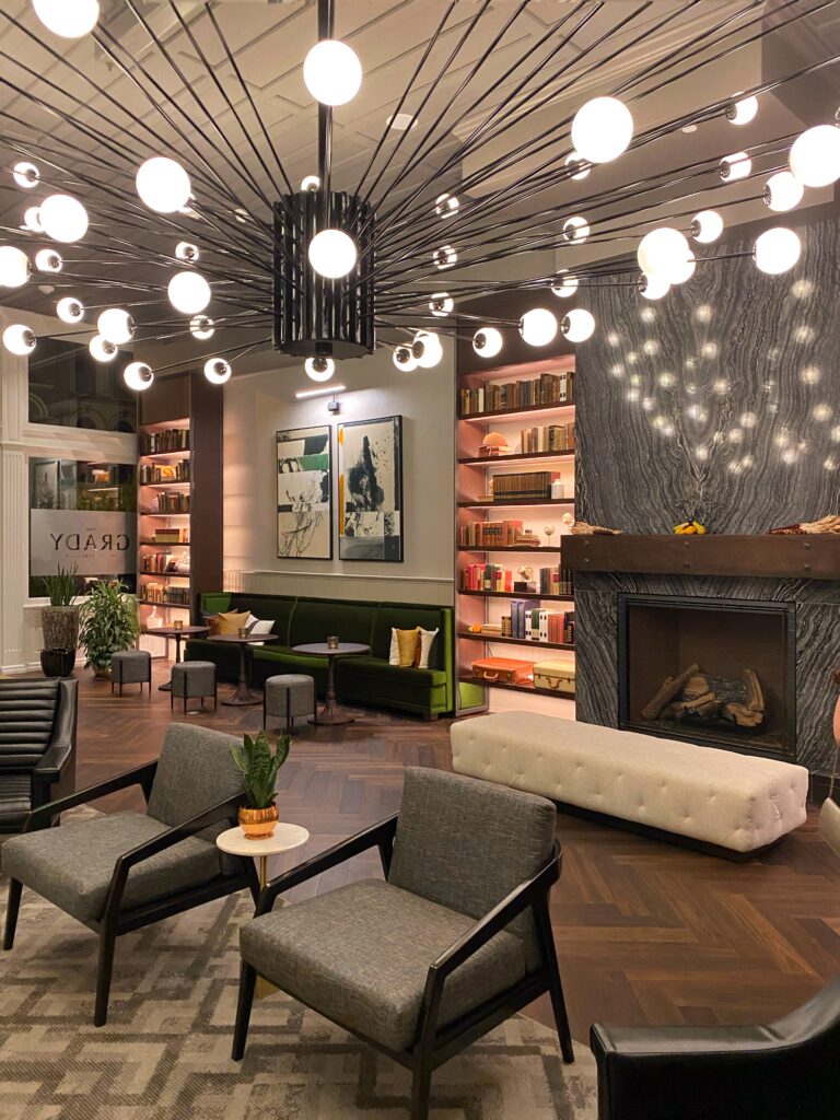 The Grady Hotel lobby in Louisville, Kentucky with a dramatic starburst chandelier, built-in bookcase shelves, green banquette seating, and a modern fireplace—an aesthetic boutique hotel lobby in downtown Louisville.