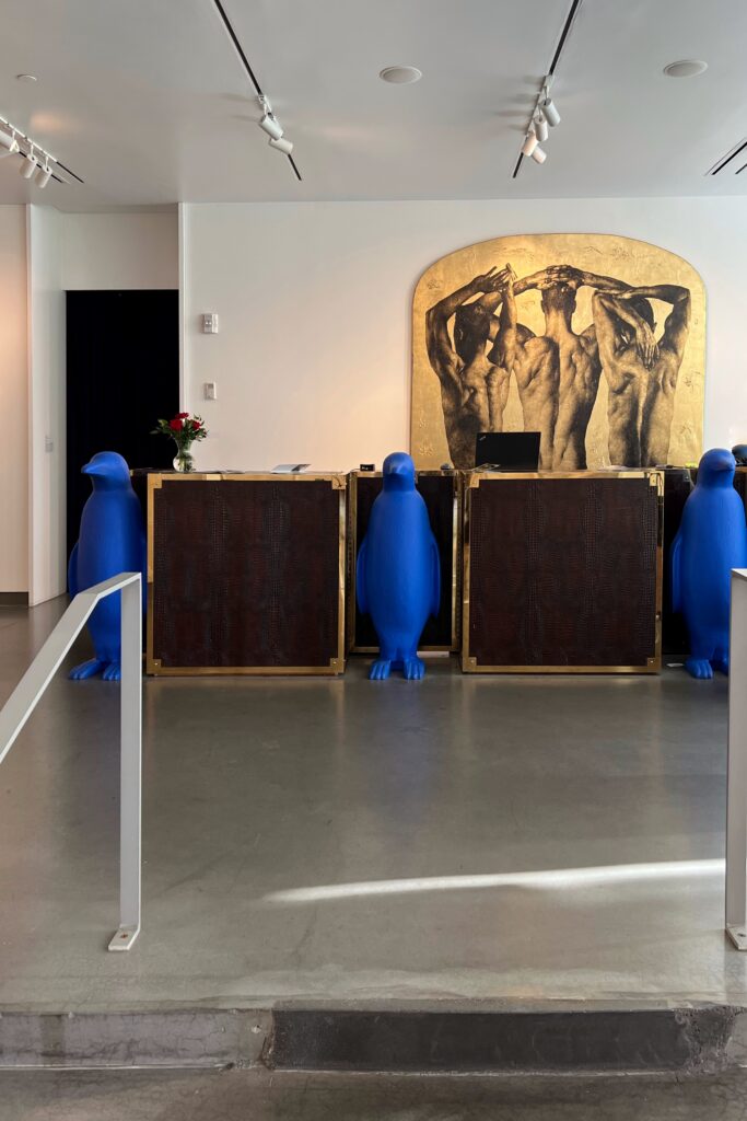 The check-in desk at 21c Museum Hotel Lexington features the brand’s signature blue penguin sculptures flanking the front counters. Behind the desk, a large gold-toned artwork adds a museum-like feel to the minimalist hotel lobby in Lexington, Kentucky.