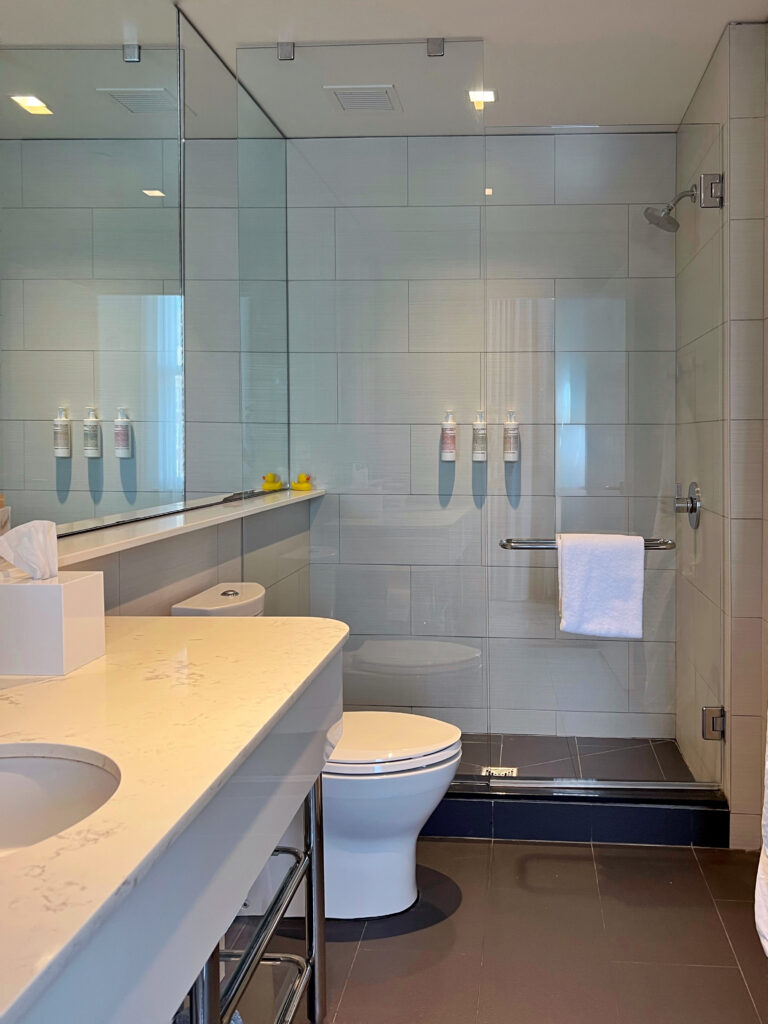 Bright, modern bathroom at 21c Museum Hotel Lexington with a glass walk-in shower, white subway tile, and dark gray floor tile. A long vanity mirror reflects wall-mounted toiletries, a towel bar, and two small yellow rubber ducks on the shower ledge.