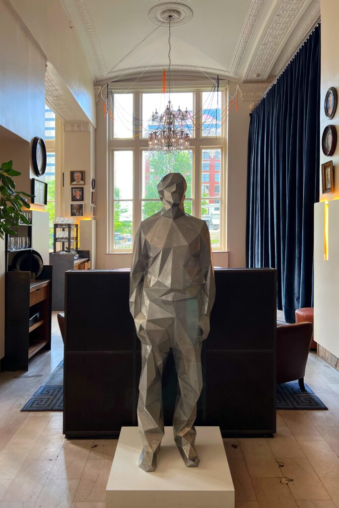 A faceted silver human sculpture stands on a white pedestal in the lobby of 21c Museum Hotel Lexington, Kentucky, framed by tall windows and dark velvet curtains. Overhead, a chandelier and draped strands of syringes add to the gallery-like atmosphere in this boutique museum hotel.
