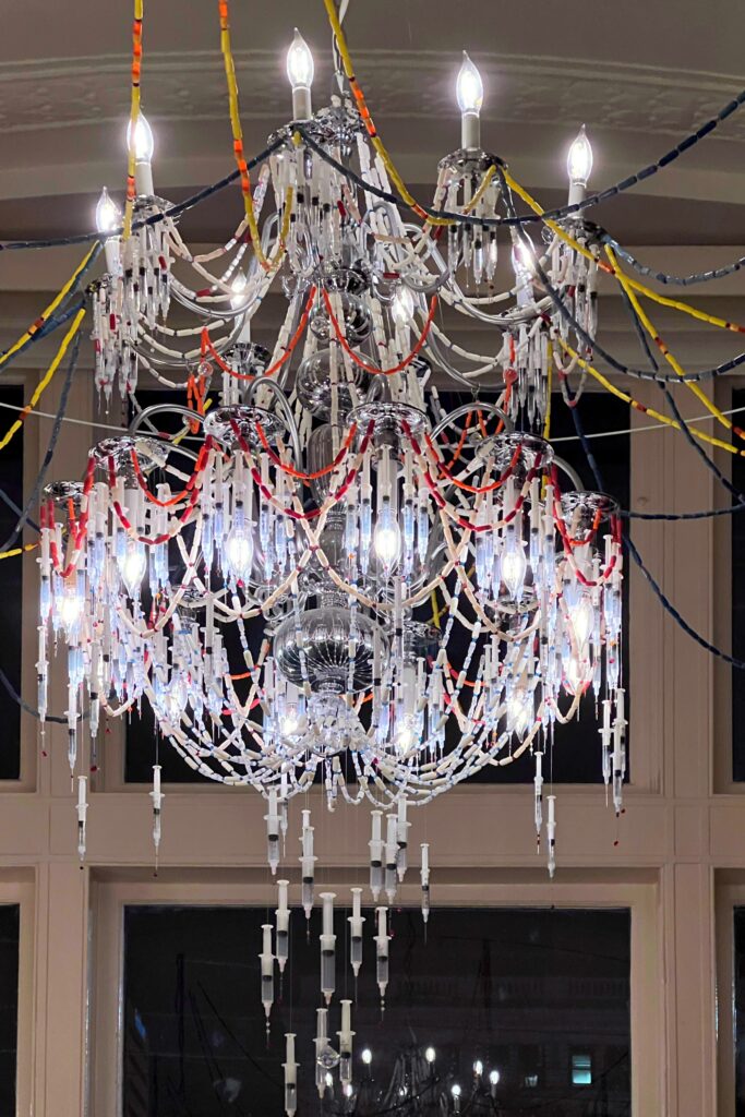 A dramatic chandelier made from clear plastic syringes hangs in the lobby at 21c Museum Hotel in Lexington, Kentucky. The sculptural fixture combines candle-style bulbs with strands of syringes and colorful tubing, creating a bold, edgy, contemporary art moment overhead.