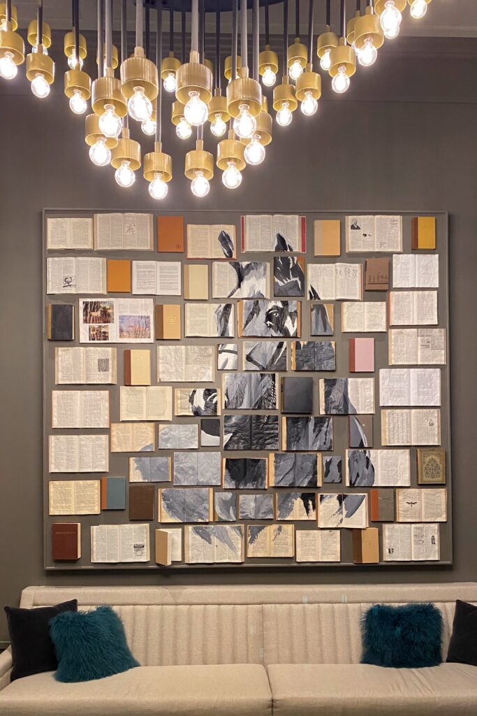 Mixed-media wall art at The Grady Hotel in Louisville features a horse image painted across a grid of vintage open books and old hardcovers. The installation hangs above a cream sofa and beneath a dramatic cluster chandelier, adding a literary, equestrian touch to the hotel’s interior design.