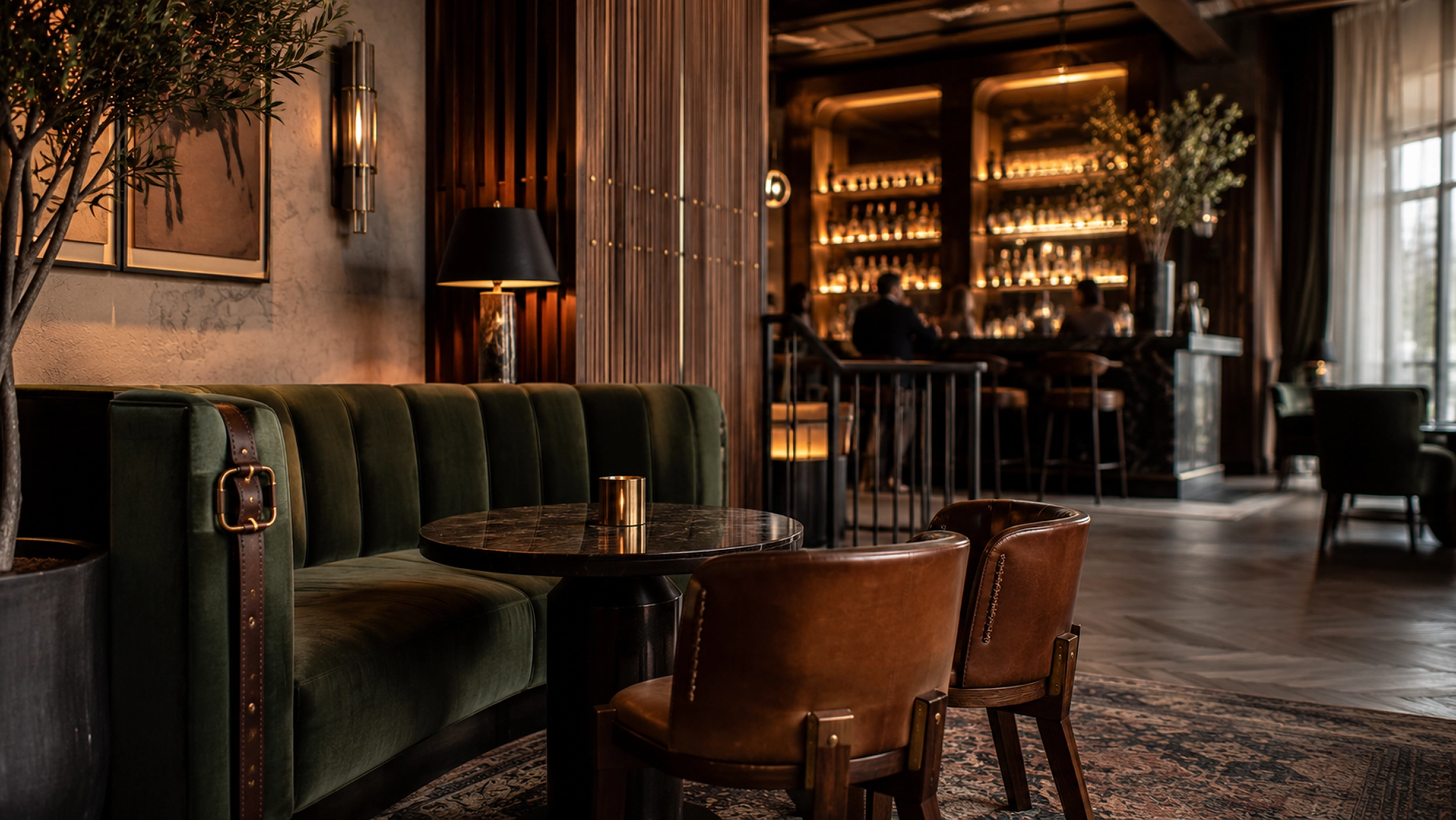 Moody hotel bar lounge with a deep green velvet banquette, round marble table, and caramel leather chairs on a patterned rug. Warm backlit shelves glow behind the bourbon bar in the distance, creating an intimate, upscale atmosphere.