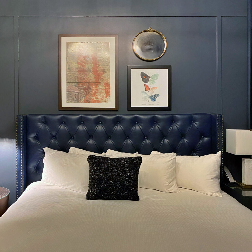Guestroom at The Grady Hotel in Lexington, Kentucky featuring a navy tufted headboard, crisp white bedding, and a dark accent pillow. Above the bed, a gallery-style arrangement includes framed artwork, a butterfly print, and a small round gilded mirror on a deep blue wall.