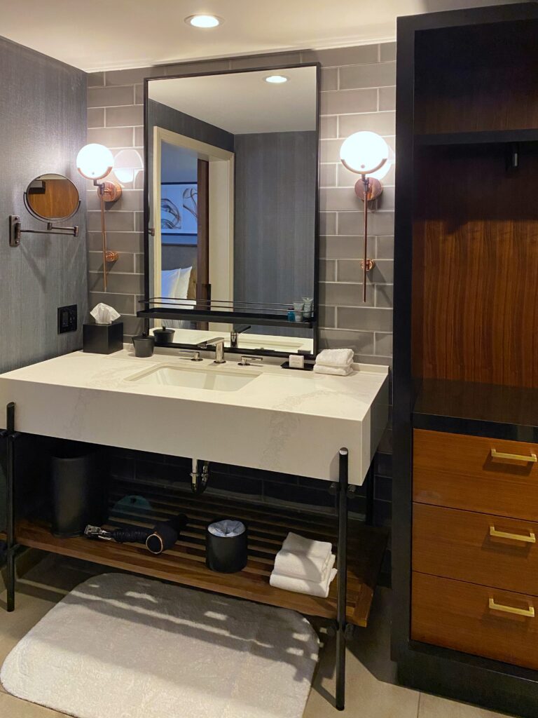 A Hotel Distil bathroom vanity features a large black-framed mirror, gray subway tile, and copper-and-glass globe sconces on both sides. Open shelving below the sink holds folded towels and amenities, adding a boutique-hotel feel.