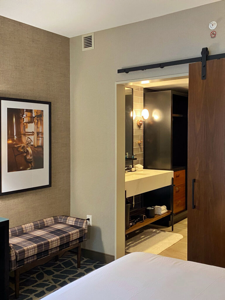 A Hotel Distil guestroom in Louisville shows a sliding wood barn door opening to a modern bathroom vanity with black cabinetry and a globe sconce. A framed distillery-inspired photo and a plaid bench add a cozy bourbon-country vibe to the room.