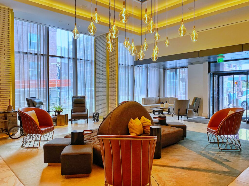 Hotel Distil’s Louisville lobby features a cluster of glowing teardrop glass pendant lights suspended over a modern seating area. Leather accent chairs and a curved sofa sit beside floor-to-ceiling windows and sheer drapes near the revolving front entrance.