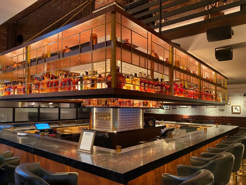 Repeal bar inside Hotel Distil in Louisville features a dramatic suspended liquor cage shelving display packed with glowing bourbon bottles and glass decanters. A dark marble bar top wraps around tufted leather seating, set against white subway tile and exposed brick accents.