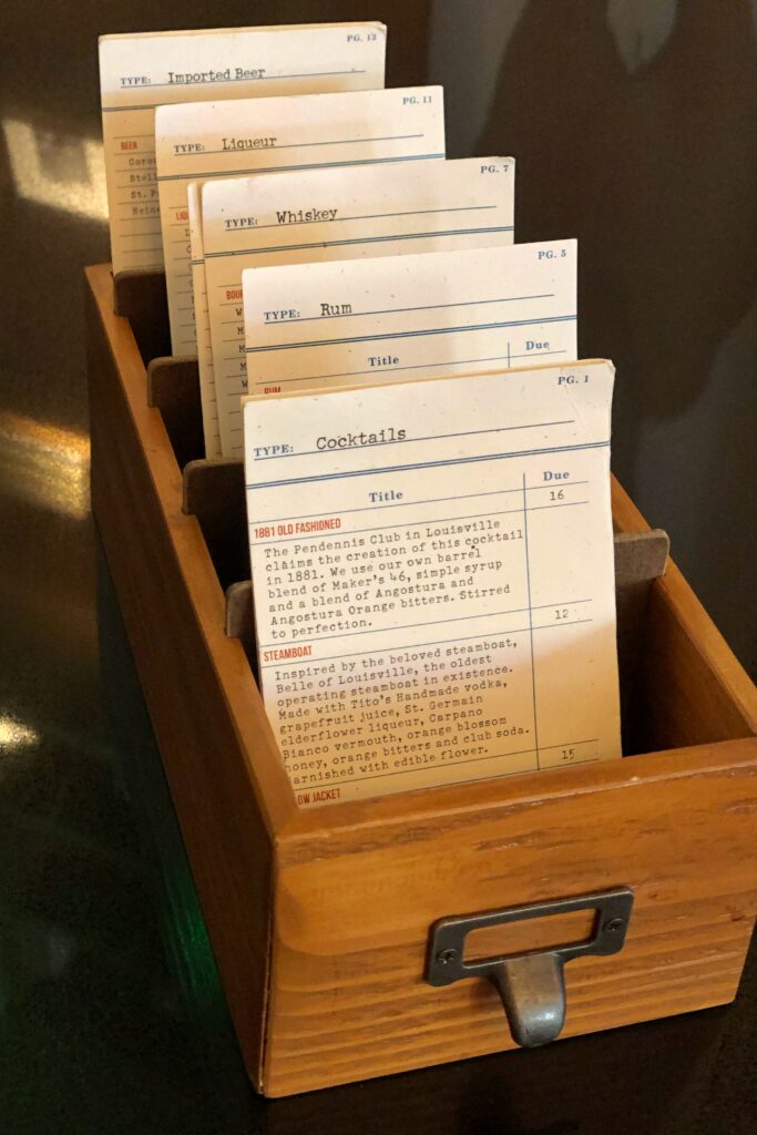 Inside the Omni Hotel Louisville Lobby Bar: a cocktail menu is displayed inside a vintage wooden card catalog drawer, with tabbed cards labeled Cocktails, Rum, Whiskey, Liqueur, and Imported Beer. The old-school library filing system gives the hotel bar a nostalgic detail featuring the 1881 Old Fashioned cocktail.