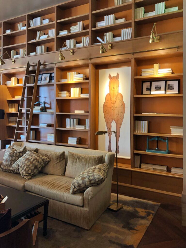 The Omni Louisville library lounge features floor-to-ceiling built-in wood bookshelves with brass picture lights and a sliding ladder. A large horse illustration panel anchors the wall above a neutral sofa, creating a warm, bourbon-country vibe in the hotel’s reading room.