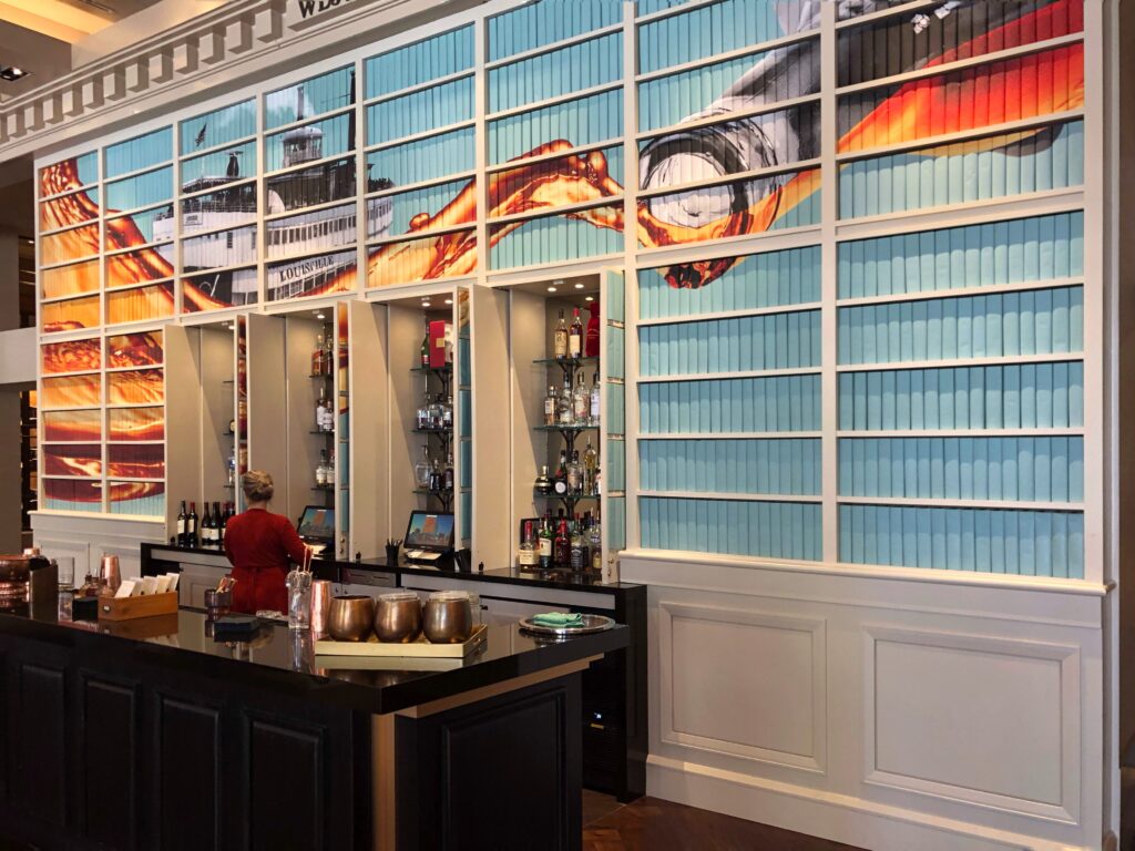 The lobby bar at Omni Louisville features a dramatic back wall with a large-scale bourbon-inspired mural stretching across the backs of books in the bookshelves. Built-in shelves display bottles alongside rows of decorative book spines, creating a polished, library-meets-bourbon-bar look.