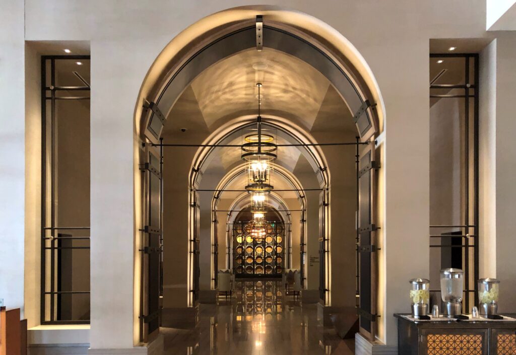 The lobby corridor at Omni Louisville Hotel features a barrel-inspired arched ceiling designed to evoke the inside of a bourbon barrel. At the far end, a dramatic wall of stacked bourbon barrels anchors the view, with warm pendant lights reflecting across the polished floor.