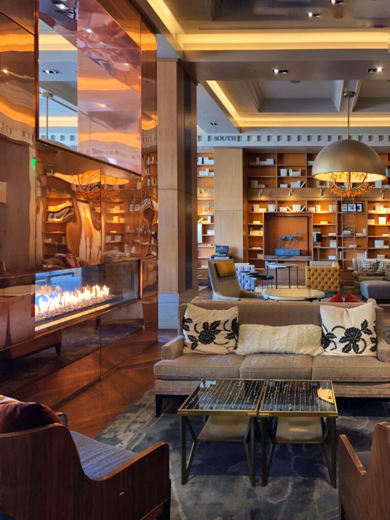 The library lounge at the Omni Hotel in Louisville features a dramatic copper-clad fireplace with a long linear flame and reflective metal panels. Floor-to-ceiling bookshelves and cozy seating create a warm, bourbon-inspired lobby vibe.
