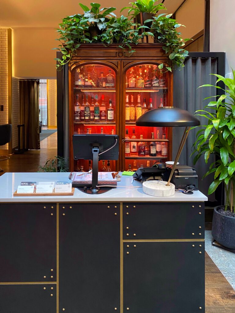 The host stand at Repeal Bar inside Hotel Distil Louisville sits in front of a glowing glass-front bourbon cabinet stocked with whiskey bottles and topped with trailing green plants. Black-and-brass cabinetry and warm red backlighting give the lobby bar a moody, bourbon-forward vibe.