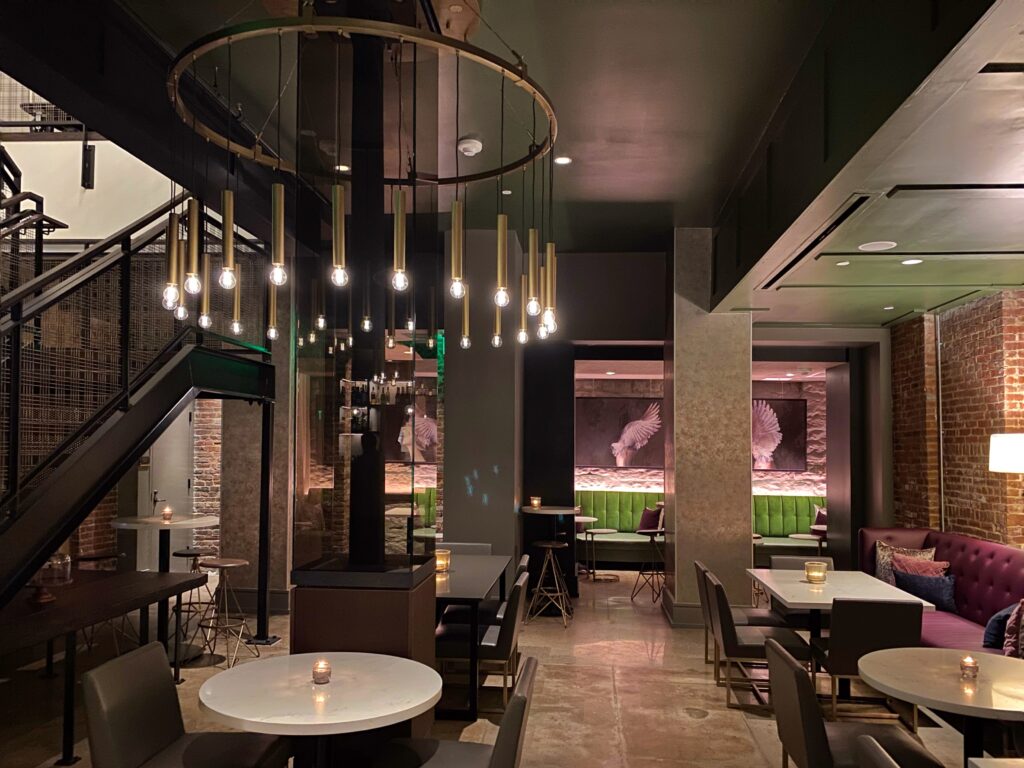 Wild Swann restaurant at The Grady Hotel in Louisville features an industrial-chic dining room with exposed brick, a black ceiling, and a circular ring of hanging Edison bulbs. Green velvet banquettes and mauve tufted seating sit beneath framed wing artwork, creating a moody, design-forward atmosphere.