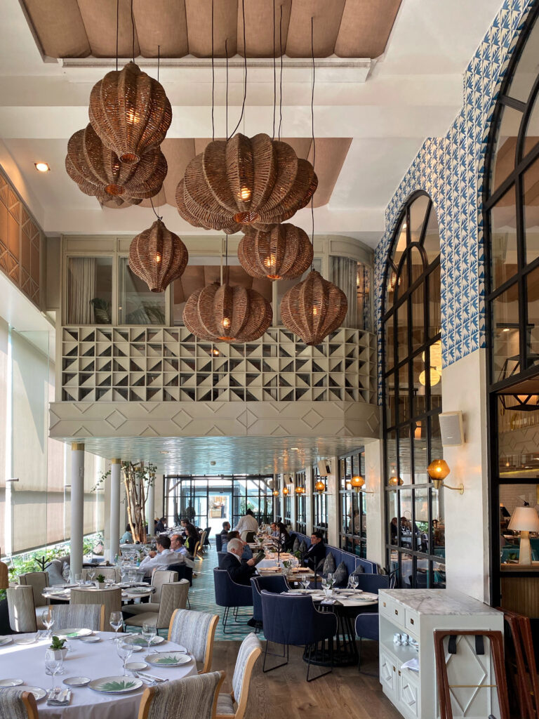 Mediterranean style dining room at Aitana in Mexico City with oversized woven pendant lights, arched black-framed windows, blue patterned tile, and layered coastal-inspired details. The airy, aesthetic restaurant interior mixes warm wood, marble, brass accents, and lush architectural texture for an elevated Mexico City dining experience.