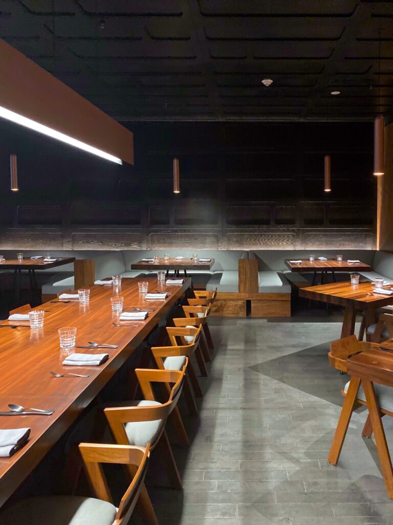 Dining room at Bajel in Mexico City with long wood tables, matching wood chairs, pale upholstered banquettes, and a dark coffered ceiling. The moody interior uses warm pendant lighting, black architectural detailing, and clean table settings for a polished, modern Mexico City restaurant atmosphere.
