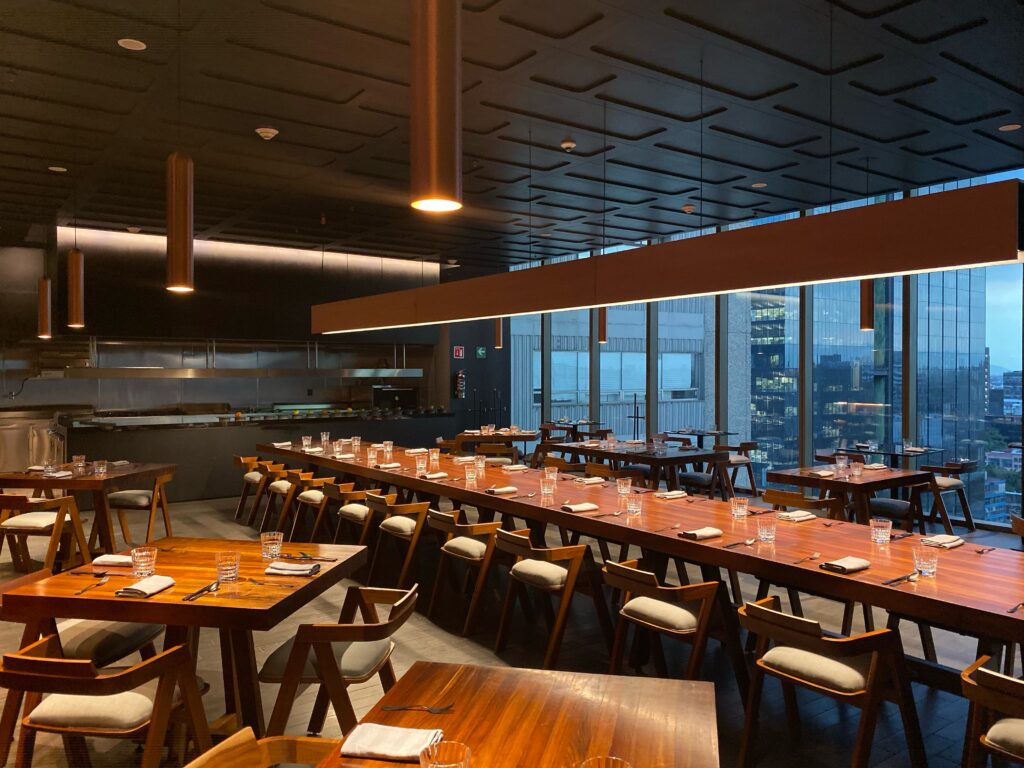 Dining room at Bajel in Mexico City with warm wood tables, modern pendant lighting, an open kitchen, and floor-to-ceiling windows overlooking the city skyline. The polished interior pairs clean architectural lines with soft evening light for an elevated Mexico City restaurant experience.