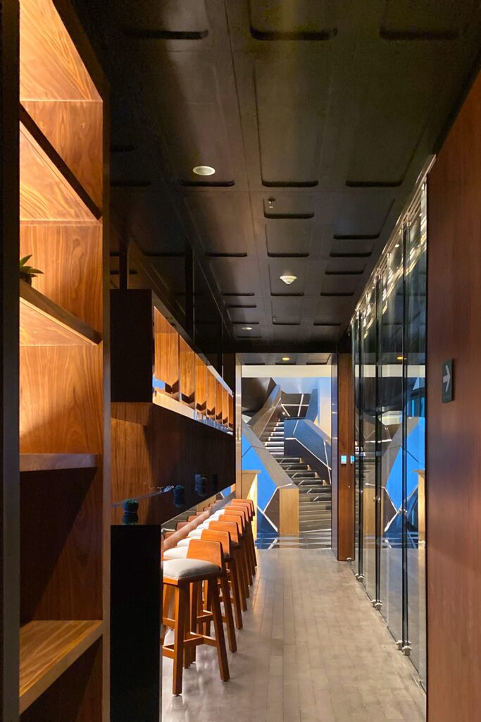 Interior hallway at the bar at Bajel in Mexico City with warm wood paneling, a row of bar stools, glass walls, and a dark coffered ceiling leading toward the lobby's sculptural staircase.